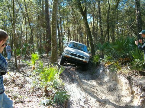 1991 Ford Bronco - A silver SUV navigates a challenging off-road trail through a dense forest.