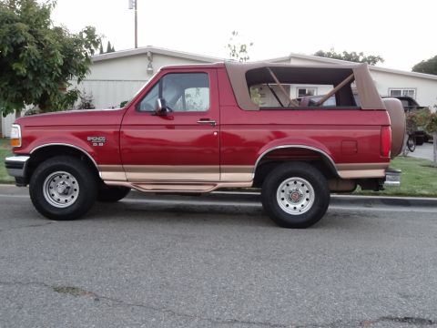 1994 Ford Bronco - This red 1994 Ford Bronco features a tan soft top and is viewed from the side, showing its chrome trim and spare tire.
