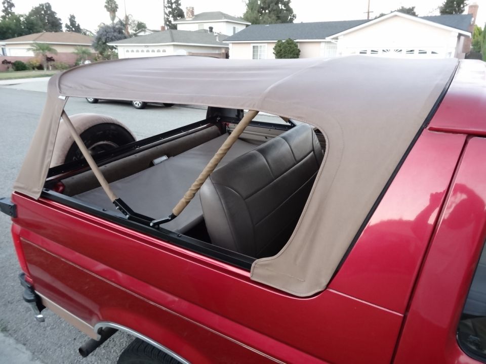 Red Ford Bronco with tan soft top, rear passenger side view. Spare tire visible