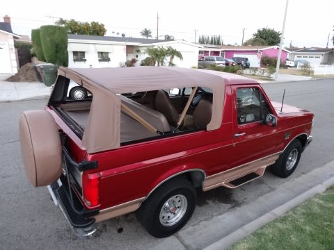 1994 Ford Bronco - This red Ford Bronco features a tan soft top and is parked curbside in a suburban neighborhood.
