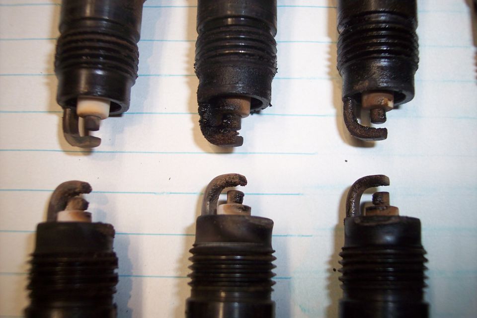 Six used spark plugs, some with heavy carbon buildup, on lined paper