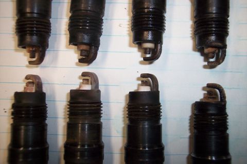 1990 Ford Bronco - A collection of eight spark plugs are displayed on a surface with a grid pattern. The condition of the electrodes varies.