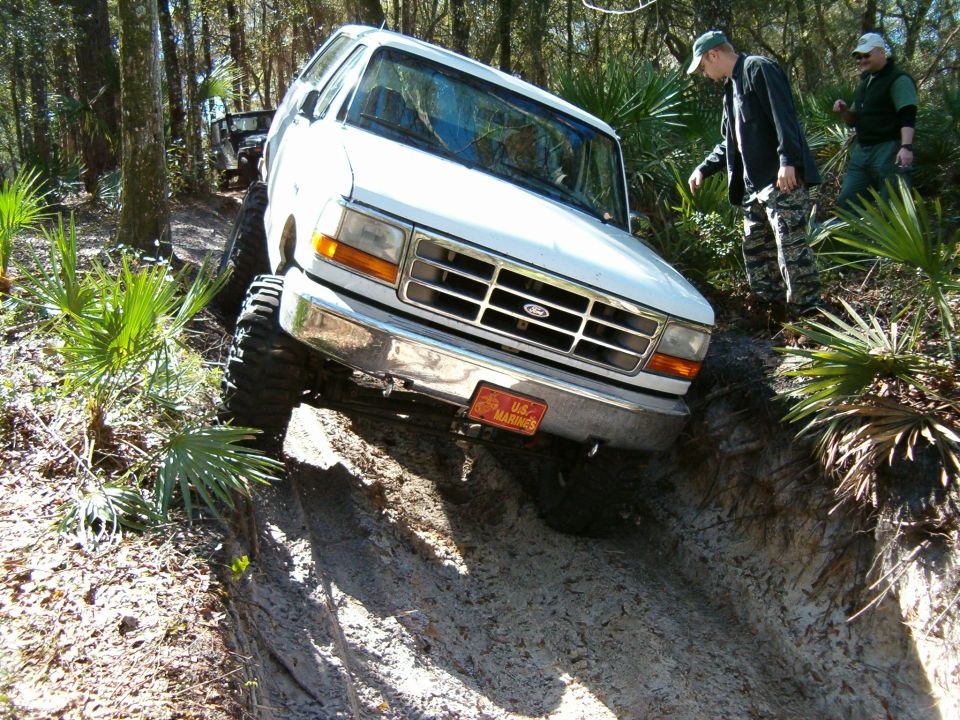 White Ford Bronco driving down a dirt path in the woods, another vehicle behind it