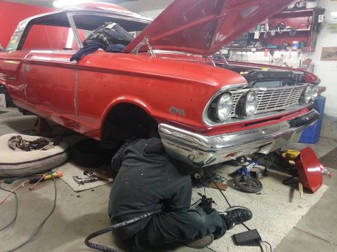 1964 Ford Fairlane - A person works on the front end of a red classic car, likely a 1964 Ford Fairlane, with its hood open. The car is raised on jack stands in…