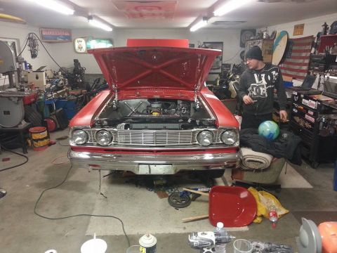 1964 Ford Fairlane - A bright red 1964 Ford Fairlane is parked in a garage with its hood open, revealing the engine bay. A man stands next to the car, looking a…