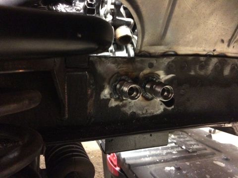 1964 Ford Fairlane - Two metal fittings are welded onto the underside of a black metal frame, likely part of a vehicle's chassis. The fittings appear to be thre…