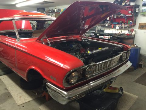 1964 Ford Fairlane - A red 1960s era Ford Fairlane is shown in a garage with its hood open, revealing the engine bay. The car is on jack stands and appears to b…