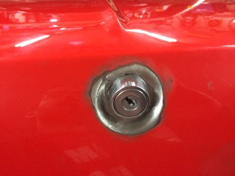 1964 Ford Fairlane - A close-up shot of a silver keyhole lock cylinder set into a bright red car body panel. The lock is surrounded by a rough, metallic mountin…