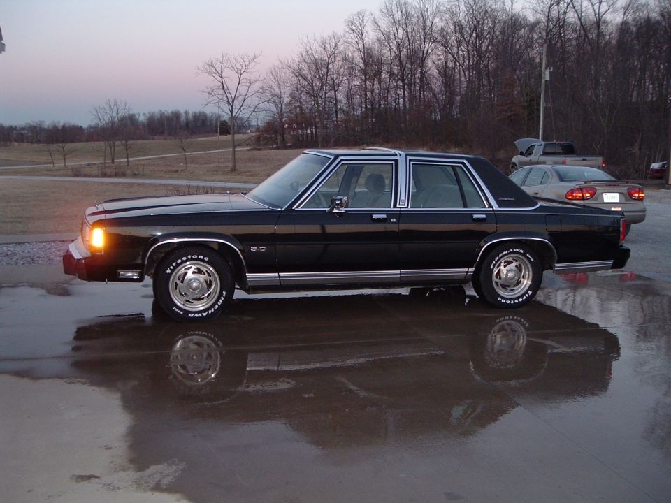 Black Ford LTD Crown Victoria sedan on white-letter tires parked on wet pavement at dusk