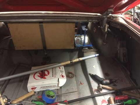 1964 Ford Fairlane - The open trunk of a red classic car reveals a custom wooden panel mounted above a fuel tank, with various tools and a box of Wilwood rotor…