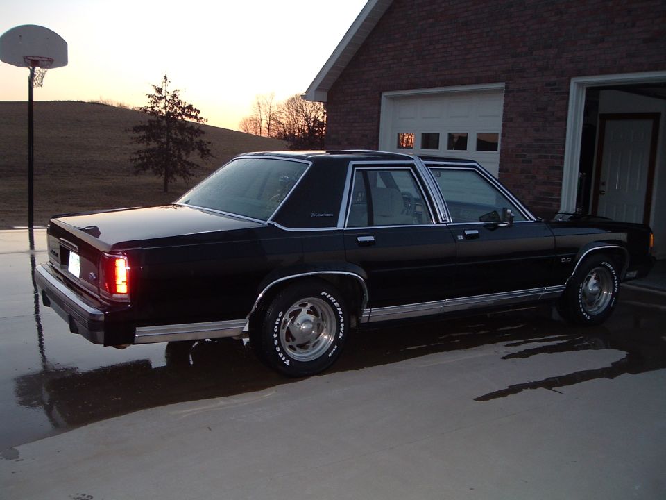 Black Ford LTD Crown Victoria sedan beside a brick garage and basketball hoop at sunset