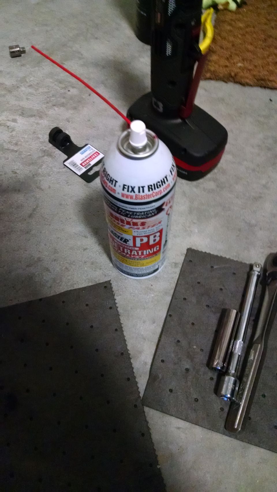 Can of PB Blaster penetrating lubricant, socket wrench, and absorbent pads on a concrete floor.