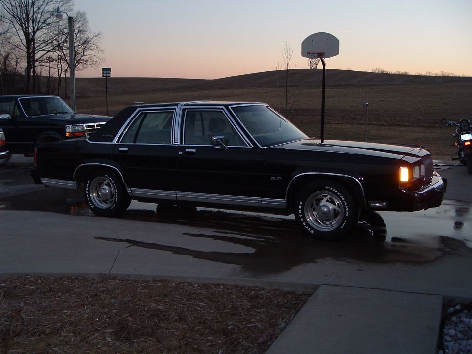Black Ford LTD sedan front three-quarter on a wet driveway with truck and motorcycle nearby