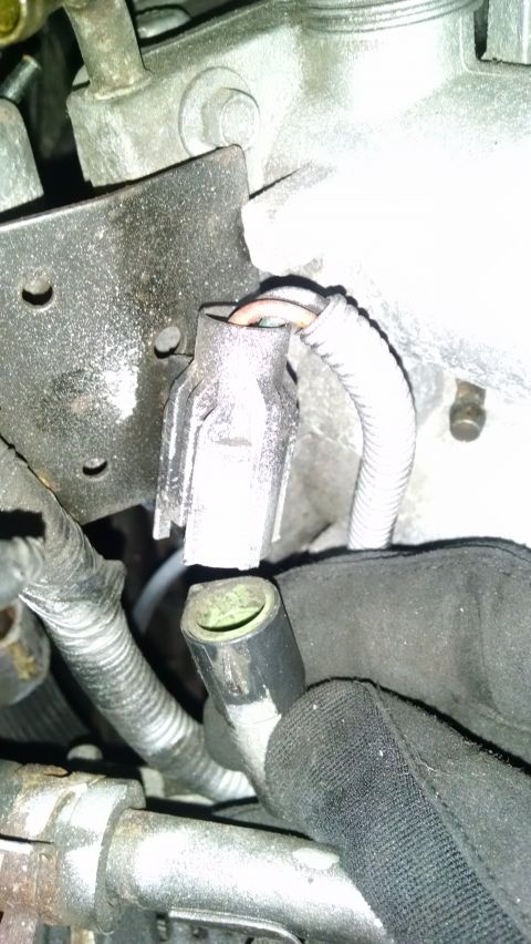 1995 Ford Bronco - A close-up view of a gray electrical connector with wires attached, situated near a metal bracket and a ribbed protective sleeve. A gloved…