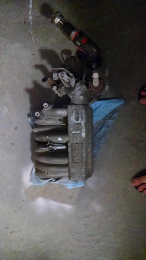 1995 Ford Bronco - A collection of car parts, including an intake manifold and a drill with a wire brush attachment, are laid out on a concrete floor.