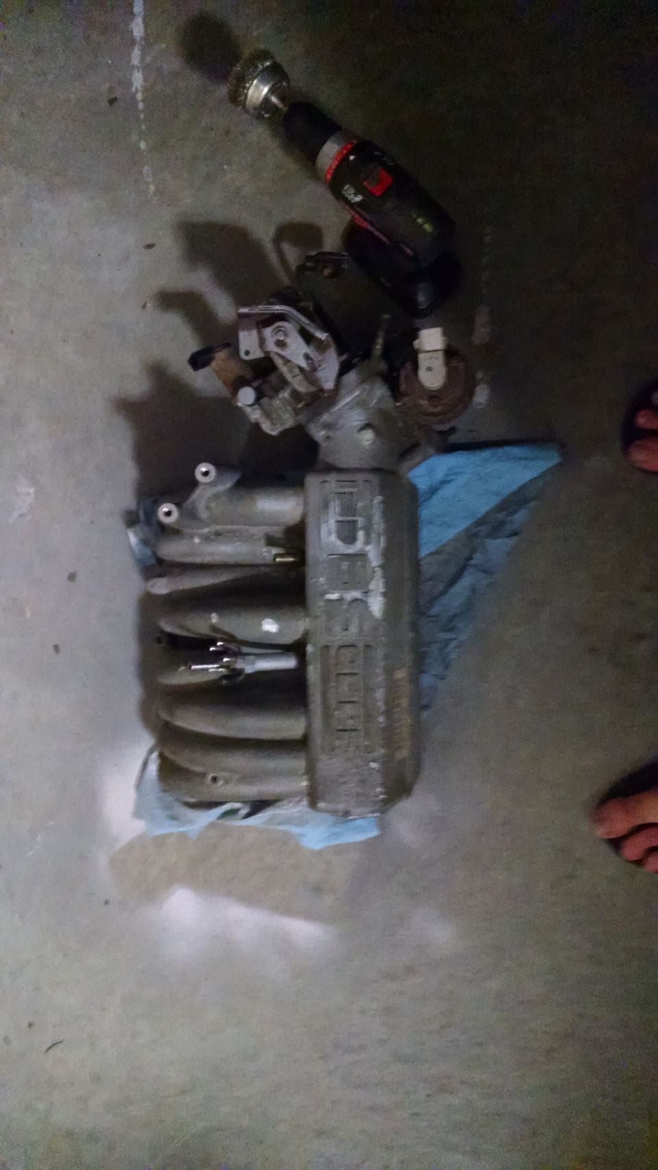 Car parts including intake manifold and drill on concrete floor.