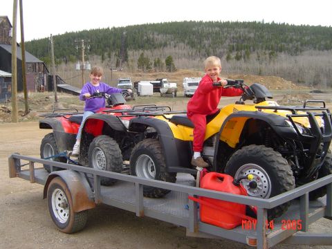1996 Ford Bronco - Two children are riding on ATVs loaded onto a trailer. The ATVs are red and yellow, and the trailer has a red gas can attached to the front.