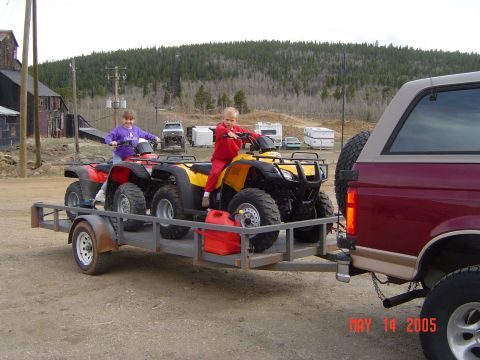 1996 Ford Bronco - Two children ride ATVs loaded onto a trailer being towed by a maroon Ford Bronco. The trailer also carries two red gas cans.