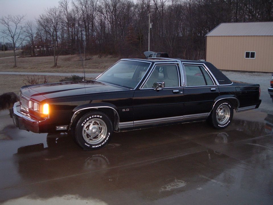 Black Ford LTD Crown Victoria sedan, headlight on, parked on wet pavement at dusk