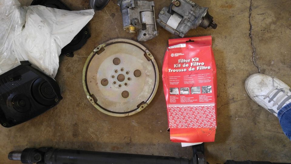 Automotive parts including flywheel, starters, filter kit, and drive shaft on concrete floor.