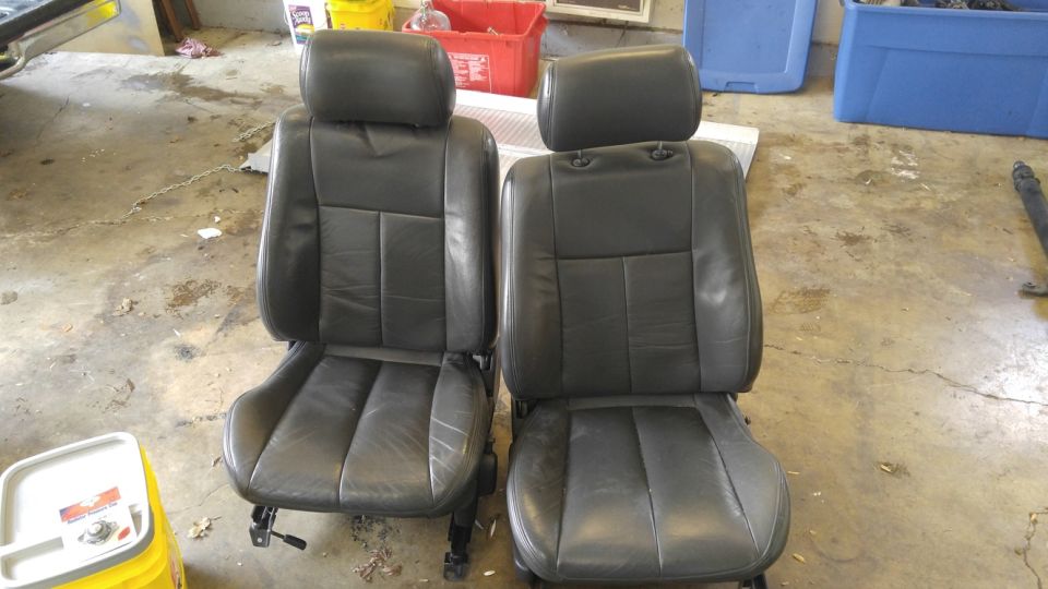 Two black leather car seats with headrests on a concrete floor.