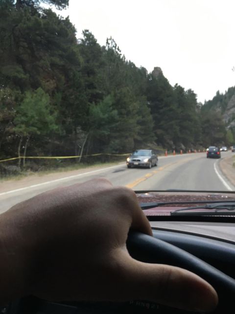 1996 Ford Bronco - A hand grips the steering wheel of a vehicle driving on a road through a forested mountain pass. Orange cones and yellow caution tape line…