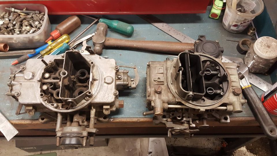 Two automotive carburetors on a workbench with tools and hardware.