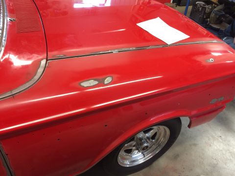 1964 Ford Fairlane - A close-up, side view of a red classic car undergoing restoration. The hood and fender show areas of bodywork, including primer and visible…