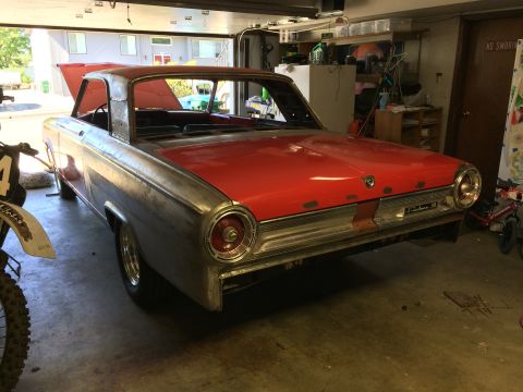 1964 Ford Fairlane - A partially restored, red and unpainted metal 1960s coupe is parked inside a garage. The car's hood is open, revealing the interior, and a…