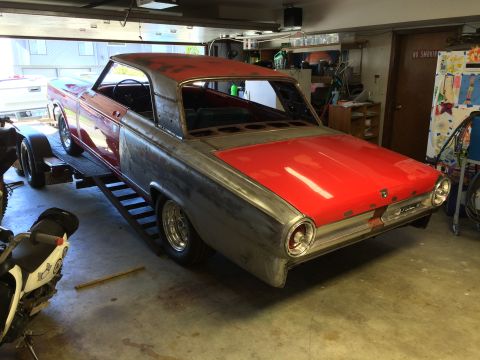 1964 Ford Fairlane - A red and silver 1964 Ford Fairlane is parked on a trailer in a garage, appearing to be in the middle of a restoration. The car has its hoo…