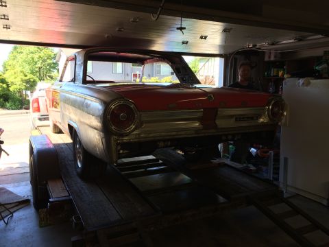1964 Ford Fairlane - A vintage red and silver car, possibly a Ford Fairlane, is parked on a trailer inside a garage. A man is visible in the driver's seat, look…