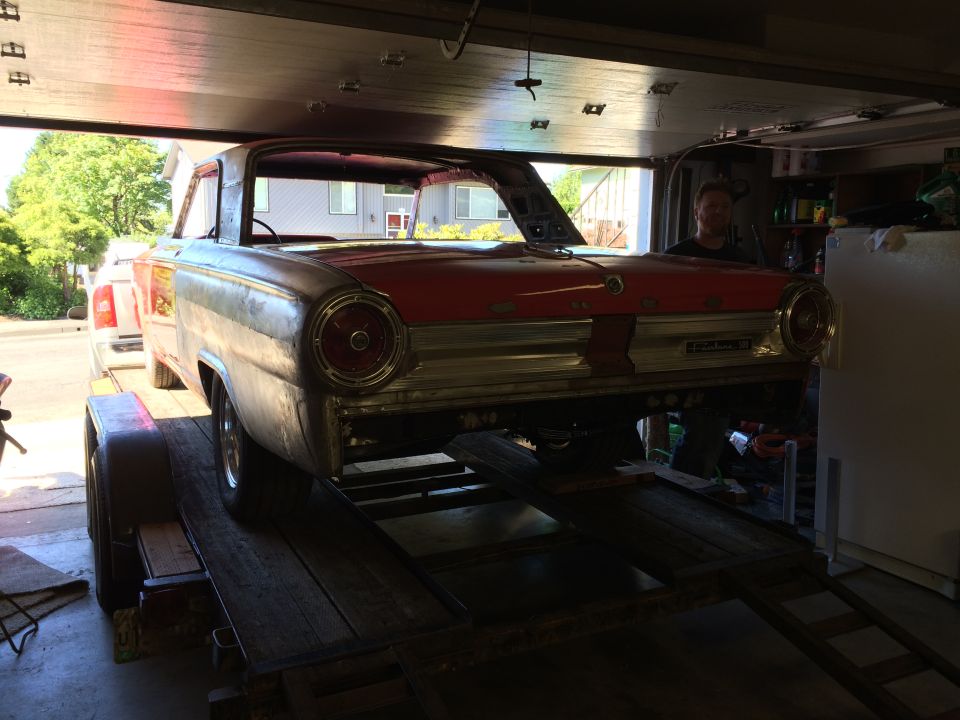 Vintage red and silver car on a trailer in a garage, with a man in the driver's seat.
