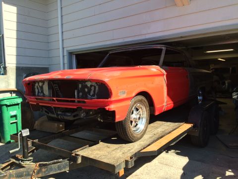 1964 Ford Fairlane - A vintage red and black car is parked on a trailer in front of an open garage door. The front of the car is missing its grille and headligh…