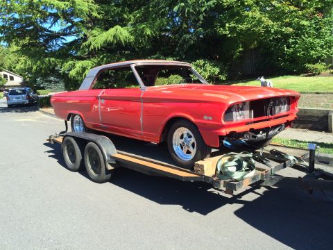 1964 Ford Fairlane - A bright red, two-door hardtop car is loaded onto a trailer. The car is missing its front grille and headlights, and appears to be undergoi…