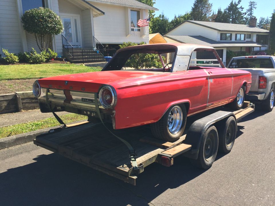 Red 1964 Ford Fairlane on a trailer, missing roof and windshield, towed by a silver pickup truck.