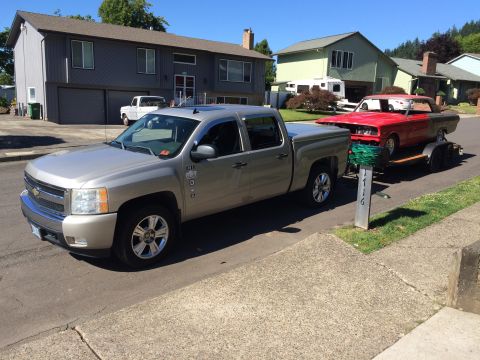1964 Ford Fairlane - A silver Chevrolet pickup truck tows a red classic car on a trailer down a suburban street. The classic car appears to be a convertible wit…