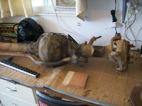 1991 Ford Bronco - A rusty Ford rear axle assembly is laid out on a workbench covered in cardboard. A black fan is visible in the background.