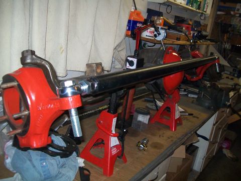 1991 Ford Bronco - A red front axle assembly with a black tie rod is mounted on red jack stands on a workbench. Various tools and parts are visible in the bac…