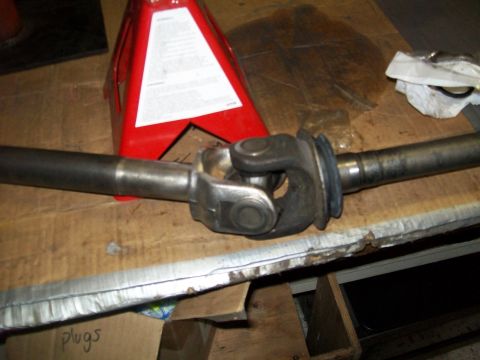 1991 Ford Bronco - A close-up view of a driveshaft with a universal joint is resting on a wooden surface. A red jack stand is visible in the background.