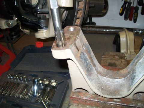 1991 Ford Bronco - A close-up shot shows a tap being used to thread a hole in a metal suspension component, likely a control arm, which is secured to a workbe…