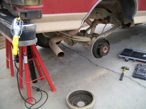 1991 Ford Bronco - The rear axle and suspension of a red and tan pickup truck or SUV are visible, supported by red jack stands. Tools and a brake drum are lai…
