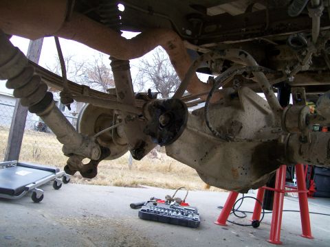 1991 Ford Bronco - The underside of a vehicle, likely a 1991 Ford Bronco, shows the rear axle, leaf springs, and shock absorber assembly. Tools and a creeper…