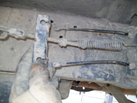 1991 Ford Bronco - A gloved hand is shown adjusting a brake cable mechanism attached to the undercarriage of a vehicle. The mechanism includes a metal bracket…
