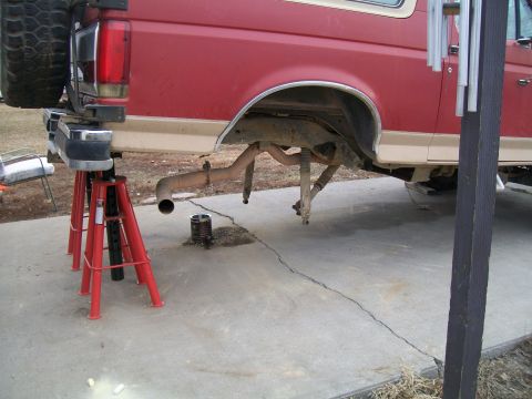 1991 Ford Bronco - The rear passenger side of a red Ford Bronco is elevated on red jack stands, with the exhaust system and rear suspension components visible…