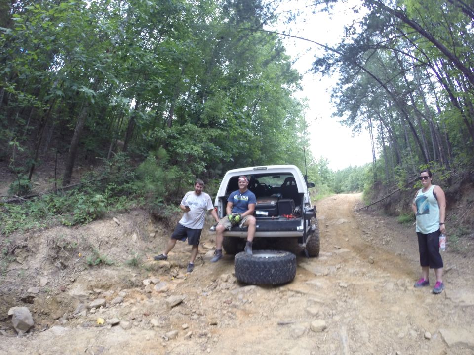 White SUV parked on a dirt road in a forest with three people standing around it.