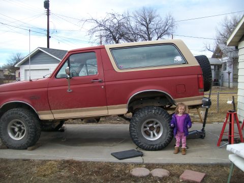 1991 Ford Bronco - A red 1991 Ford Bronco with tan trim is parked on a concrete driveway next to a house. A young girl in a purple jacket stands near the rear…