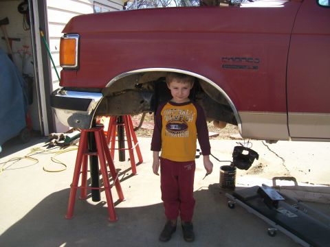 1991 Ford Bronco - A young boy stands in front of a red Ford Bronco being worked on in a garage.