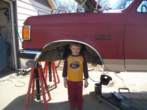 1991 Ford Bronco - A young boy stands in front of a red Ford Bronco that is raised on jack stands for maintenance.