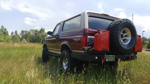 1996 Ford Bronco - The rear passenger side of a dark red 1996 Ford Bronco is shown parked in a grassy field. It features a spare tire mounted on the back, two…
