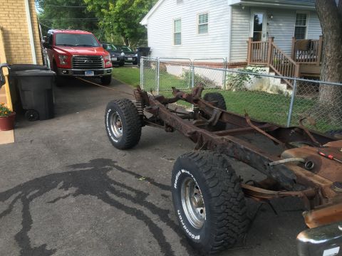1979 Ford Bronco - A rusted vehicle frame sits on pavement, with a red Ford F-150 truck in the background.